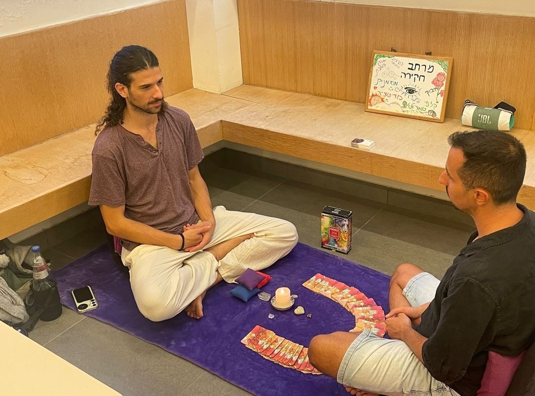 Itay Piro sitting cross-legged across from a participant, a fan of cards and a lit candle between them