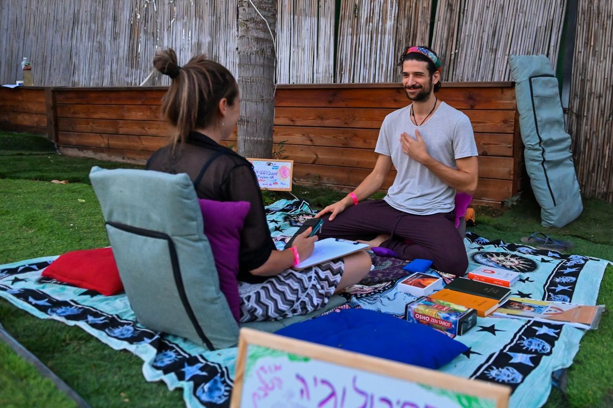 איתי פירו יושב על שמיכה בדשא מול משתתפת, עם קלפים וספרים פרוסים לצידם